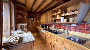 Een houten keuken met een houten aanrecht, een spoelbak en een fornuis. Er zijn planken met serviesgoed en een klok aan de muur.