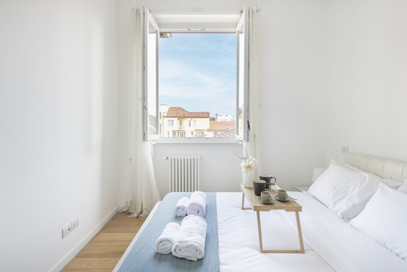 A hotel room with a bed, a small table with a cup and saucer, and a window with a view of buildings.