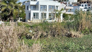 Um edifício moderno de dois andares, com grandes janelas e varanda, rodeado por vegetação e outros edifícios residenciais.