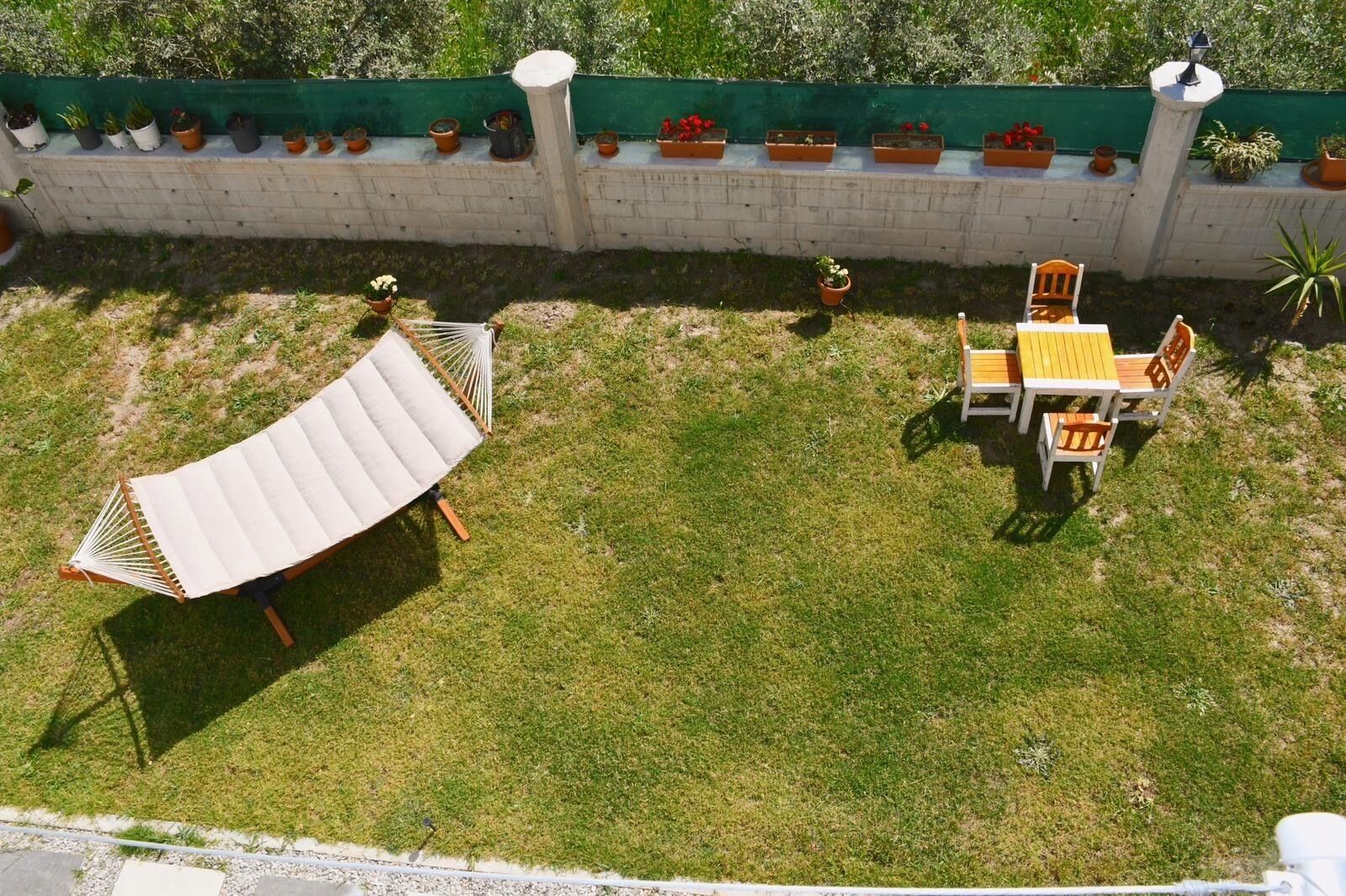 Ein grüner Außenbereich mit einer Hängematte, einem Holztisch mit Stühlen und einer Reihe von Topfpflanzen an einer weißen Mauer.