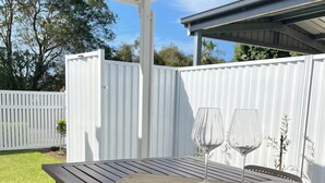 A wooden patio table with a wine glass and a wooden tray, a white fence, and a covered area with a view of trees and a building in the background.