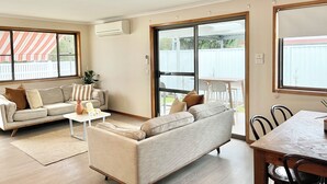 A living room with a sofa, coffee table, and dining table. There is a sliding glass door leading to an outdoor patio.