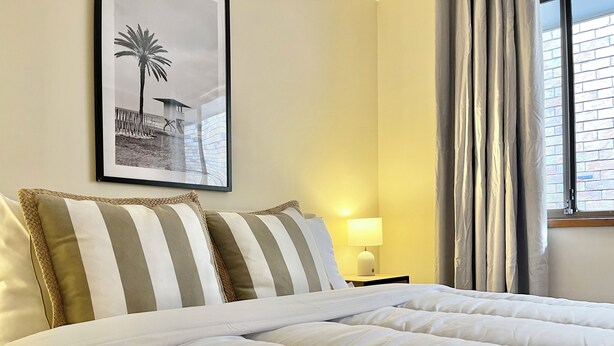 A neatly made bed with striped pillows, a framed picture on the wall, a bedside lamp, and a window with curtains.