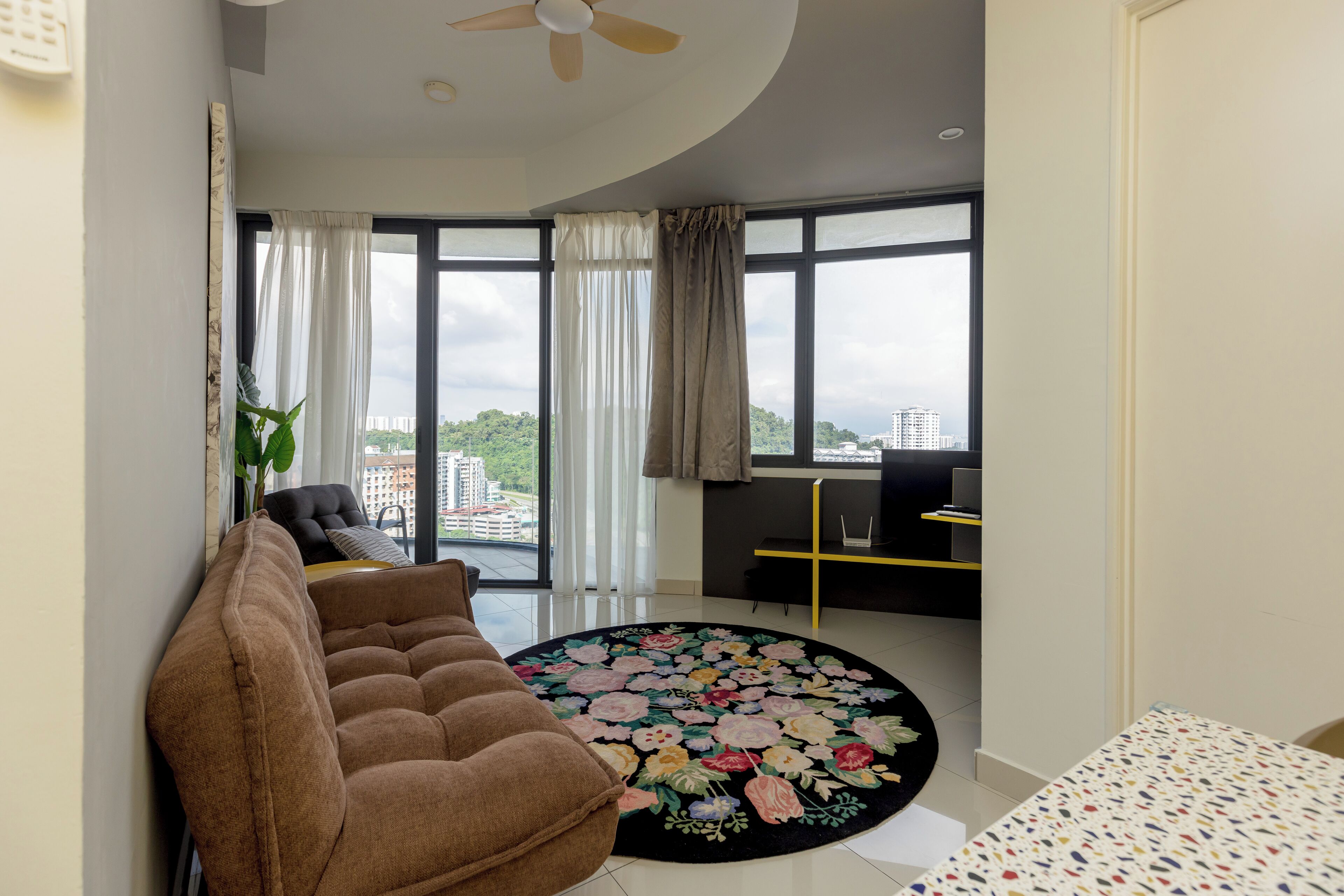 Una sala de estar moderna con un sofá marrón, un tapete redondo con motivos florales y amplios ventanales con vistas a la ciudad.