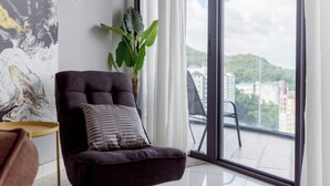 A modern living room with a dark sofa, a potted plant, and a large window offering a city view. - ArteS Suite Penang (George Town)