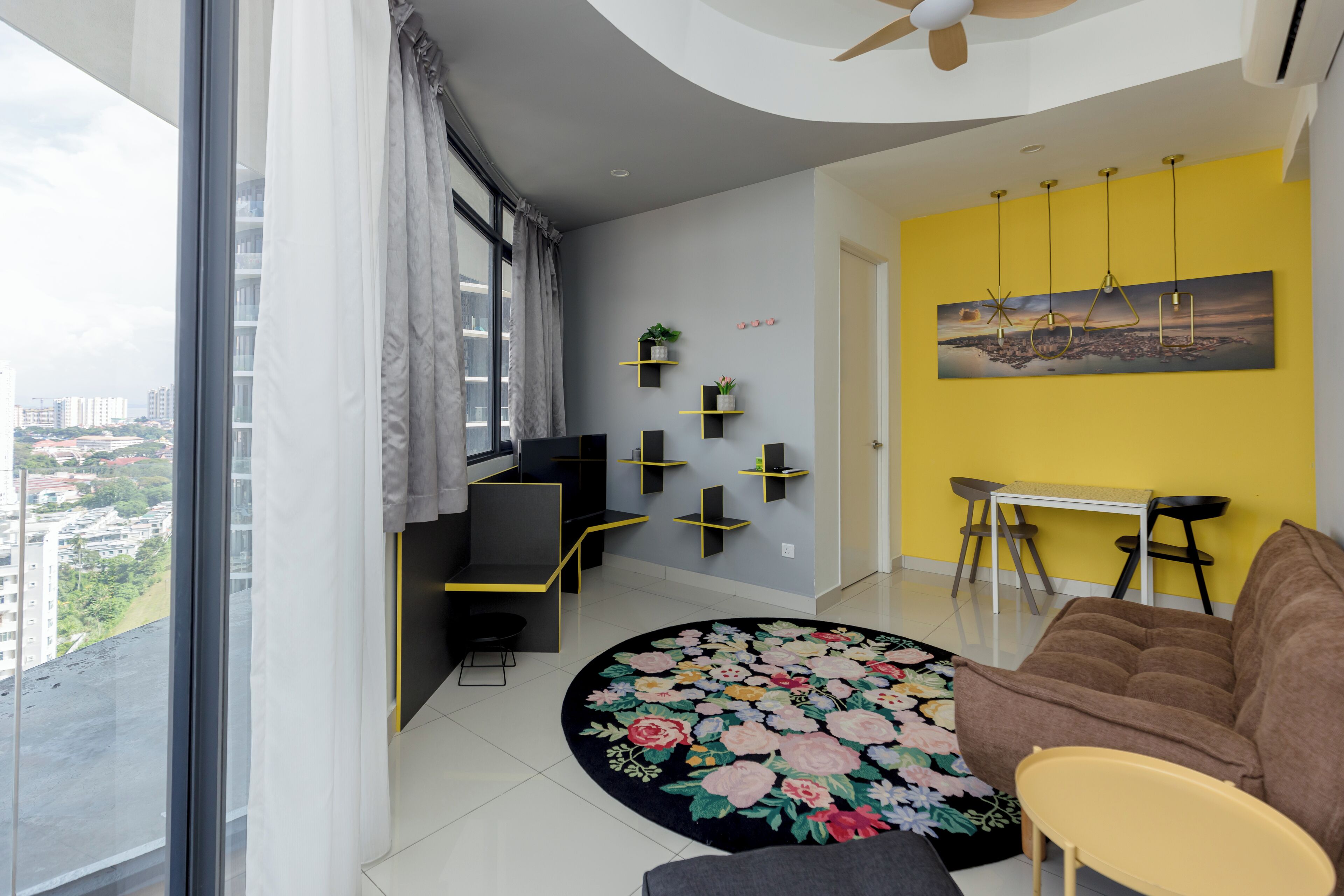 A modern living room with a large window, a round floral rug, a brown sofa, a yellow accent wall, and a city view.