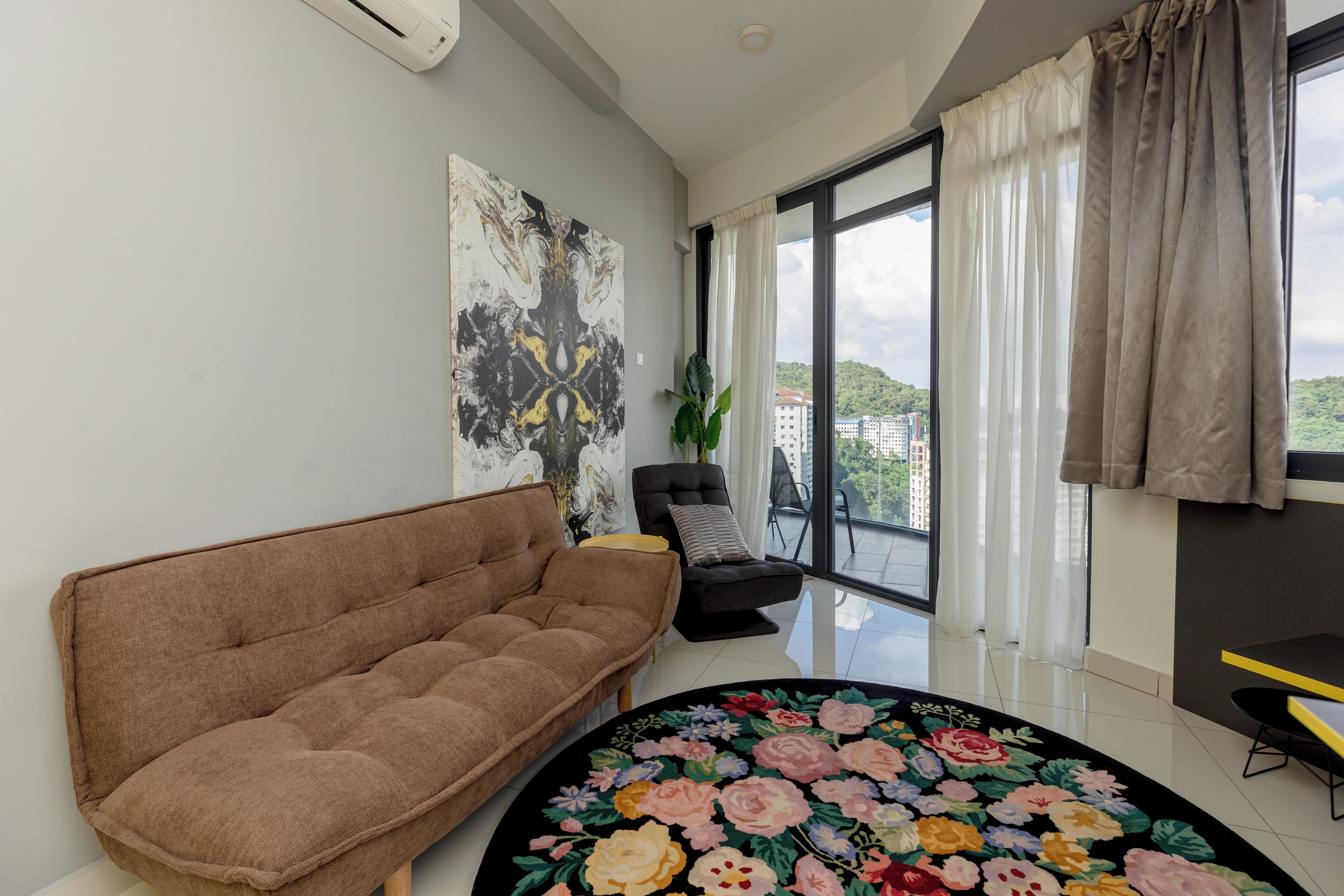 A modern living room with a brown sofa, a black armchair, a round floral rug, a large abstract painting, and a view of a cityscape through large windows.