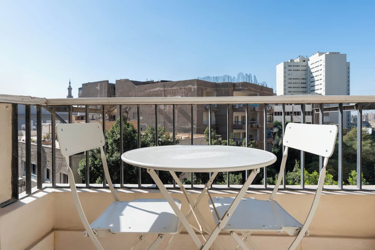 Un balcon avec une table blanche et deux chaises, donnant sur un paysage urbain composé d’immeubles et d’espaces verts.