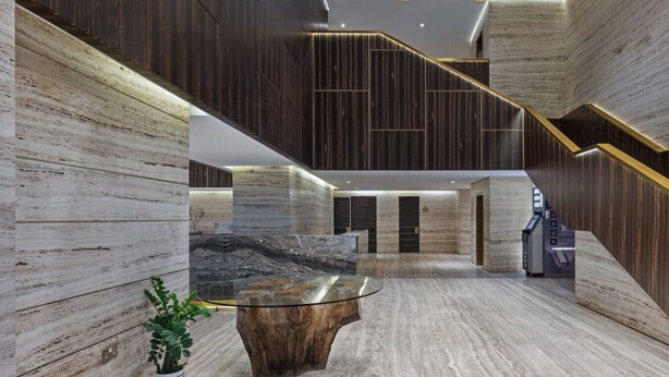Een moderne lobby met een marmeren receptiebalie, een houten salontafel en een trap met donkerhouten lambrisering.
