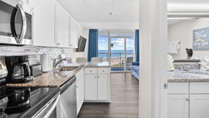 A modern kitchen with a microwave, coffee maker, and granite countertop. There is a view of the ocean through a glass door.
