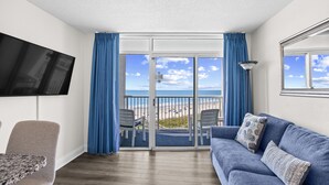 A room with a blue sofa, a TV mounted on the wall, a dining table, and a view of the beach through the sliding glass doors.