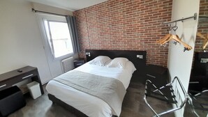 Une chambre avec un mur en briques, un lit avec une literie blanche, un bureau, une chaise et un radiateur.