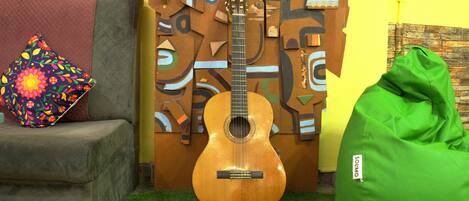 Una guitarra, un sillón de peluche verde y un cojín colorido en una habitación con una pared amarilla y un papel tapiz estampado.