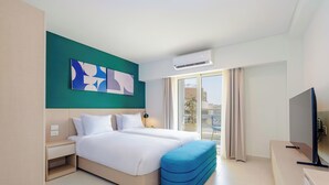 Une chambre d’hôtel moderne avec un grand lit, un pouf bleu, un téléviseur à écran plat et une vue sur la ville par la fenêtre.