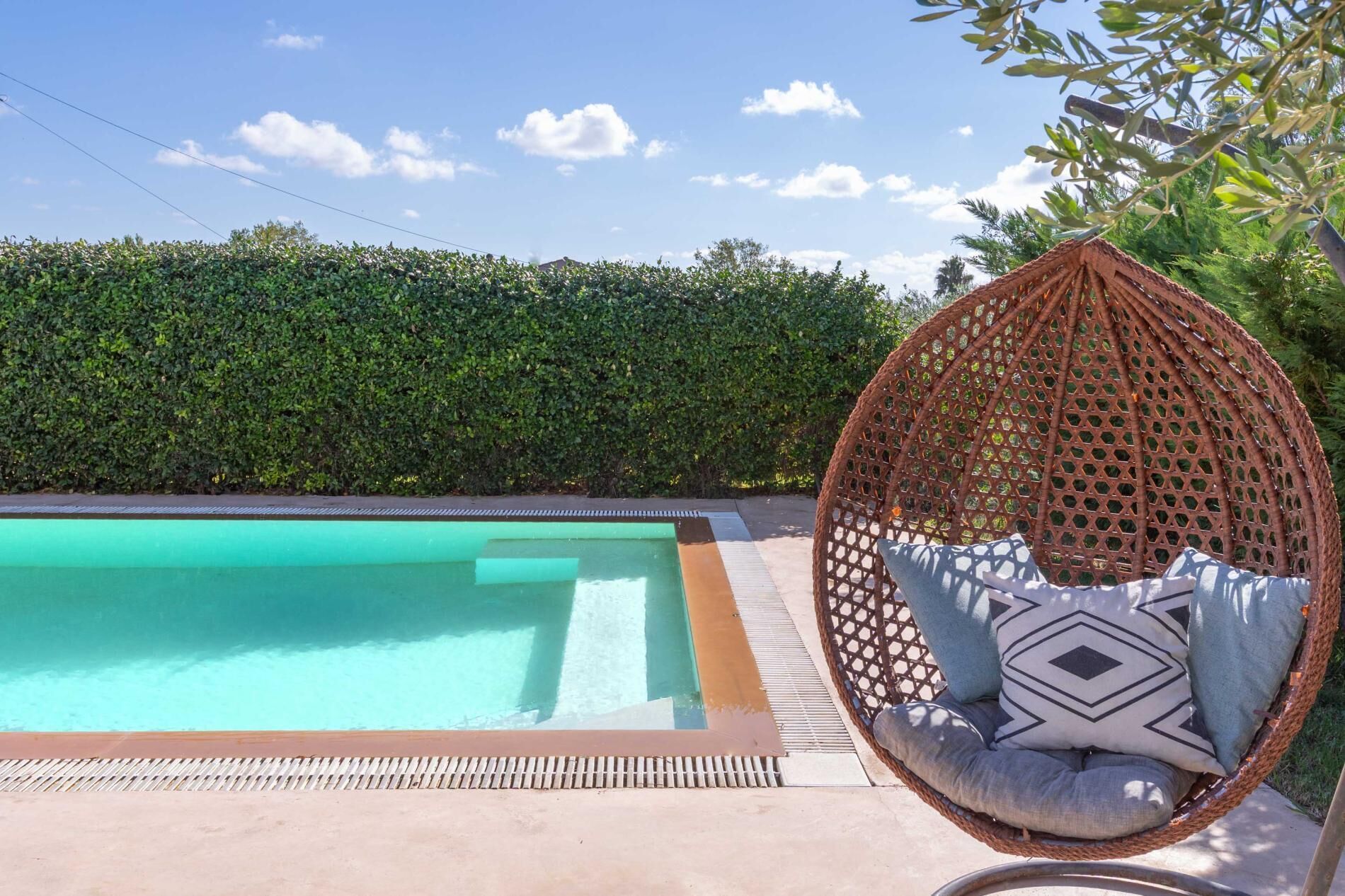 Una piscina con borde de agua azul cristalina, una silla colgante de mimbre con cojines, y un seto cuidado en el fondo.