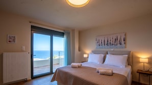 Une chambre avec un grand lit, une vue sur la mer par une porte coulissante et un tableau représentant des voiliers au mur.