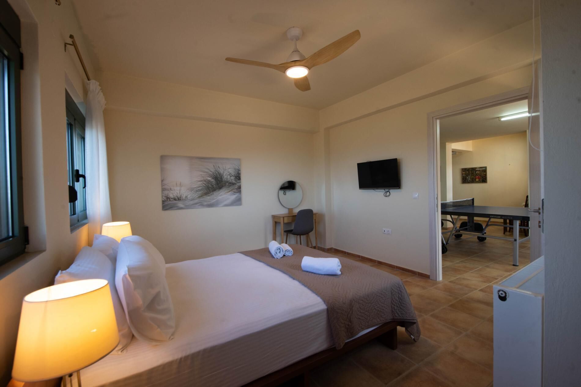 A hotel room with a bed, bedside lamps, a ceiling fan, a painting, a desk with a chair, a television, and a table with chairs.