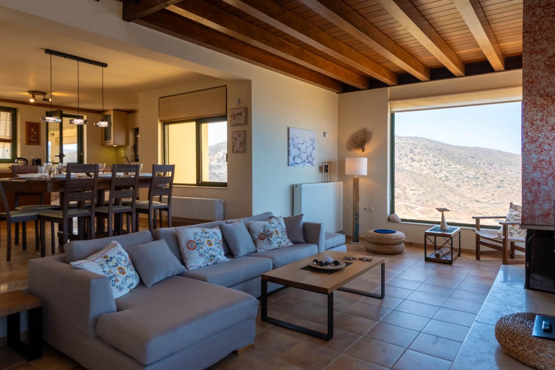 Uma sala de estar moderna com sofá, mesa de centro e área de jantar. Há uma janela grande com vista para as montanhas.