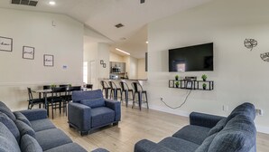 Een moderne woonkamer met grijze banken, een salontafel en een wandgemonteerde flatscreen televisie. Er is een eethoek met een tafel en stoelen, en een keuken op de achtergrond.