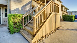 A wooden staircase leading to a porch with a railing, surrounded by greenery and a paved driveway. - Steps to Sand: Breezy Port Aransas Condo! (Port Aransas)