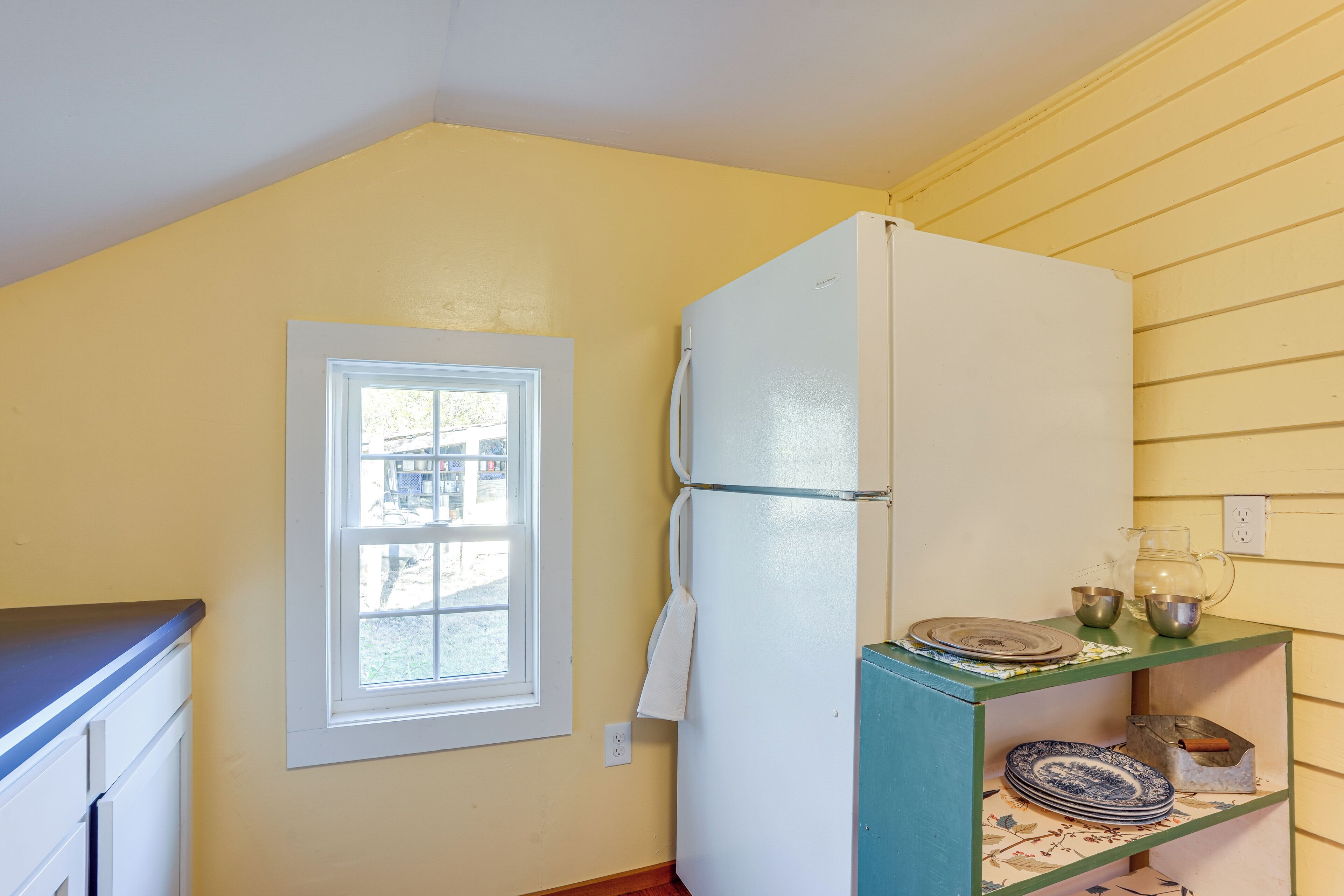 A kitchen with a white refrigerator, a window, and a shelf with dishes.