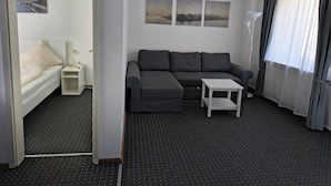 A modern hotel room with a gray sofa, a small white table, and a bed with a white headboard. There are three framed pictures on the wall and a floor lamp. - Hotel Restaurant Adria Kroatien (Bad Ems)