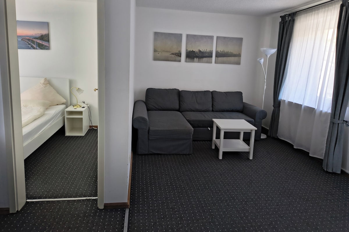 Een moderne hotelkamer met een grijze bank, een witte bijzettafeltje en een bed met een witte hoofdbord. Er hangen drie ingelijste foto’s aan de muur en staat een vloerlamp.