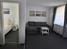 Una habitación de hotel moderna con un sofá gris, una pequeña mesa blanca y una cama con cabecera blanca. Hay tres cuadros enmarcados en la pared y una lámpara de pie.
