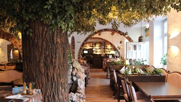 A restaurant with a rustic interior, featuring a large tree trunk, brick walls, wooden tables, and a brick archway leading to the bar area. - Hotel Restaurant Adria Kroatien (Bad Ems)