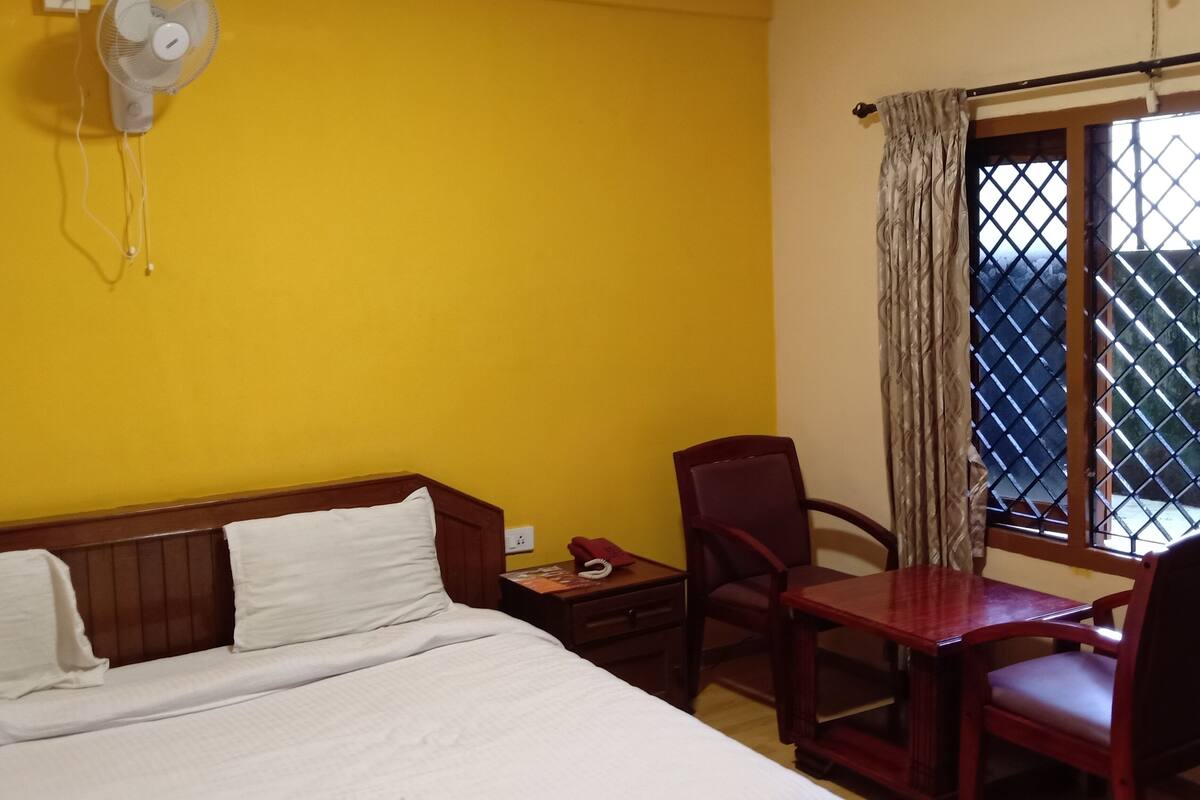Una habitación de hotel con cama, escritorio, silla, ventilador y ventana con cortinas.