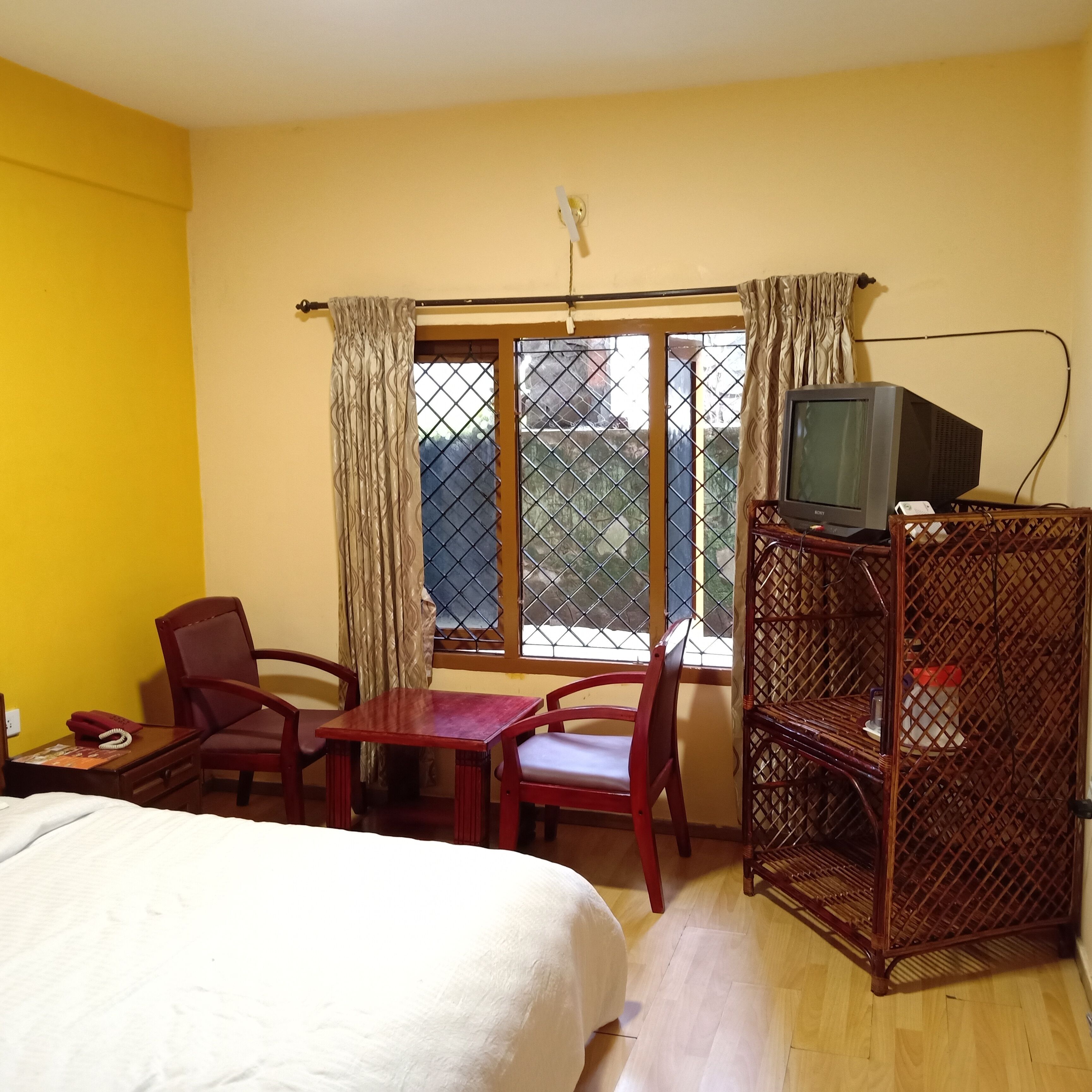 Una habitación de hotel con una cama, un escritorio con teléfono, una silla, un televisor sobre un mueble de madera y una ventana con cortinas.