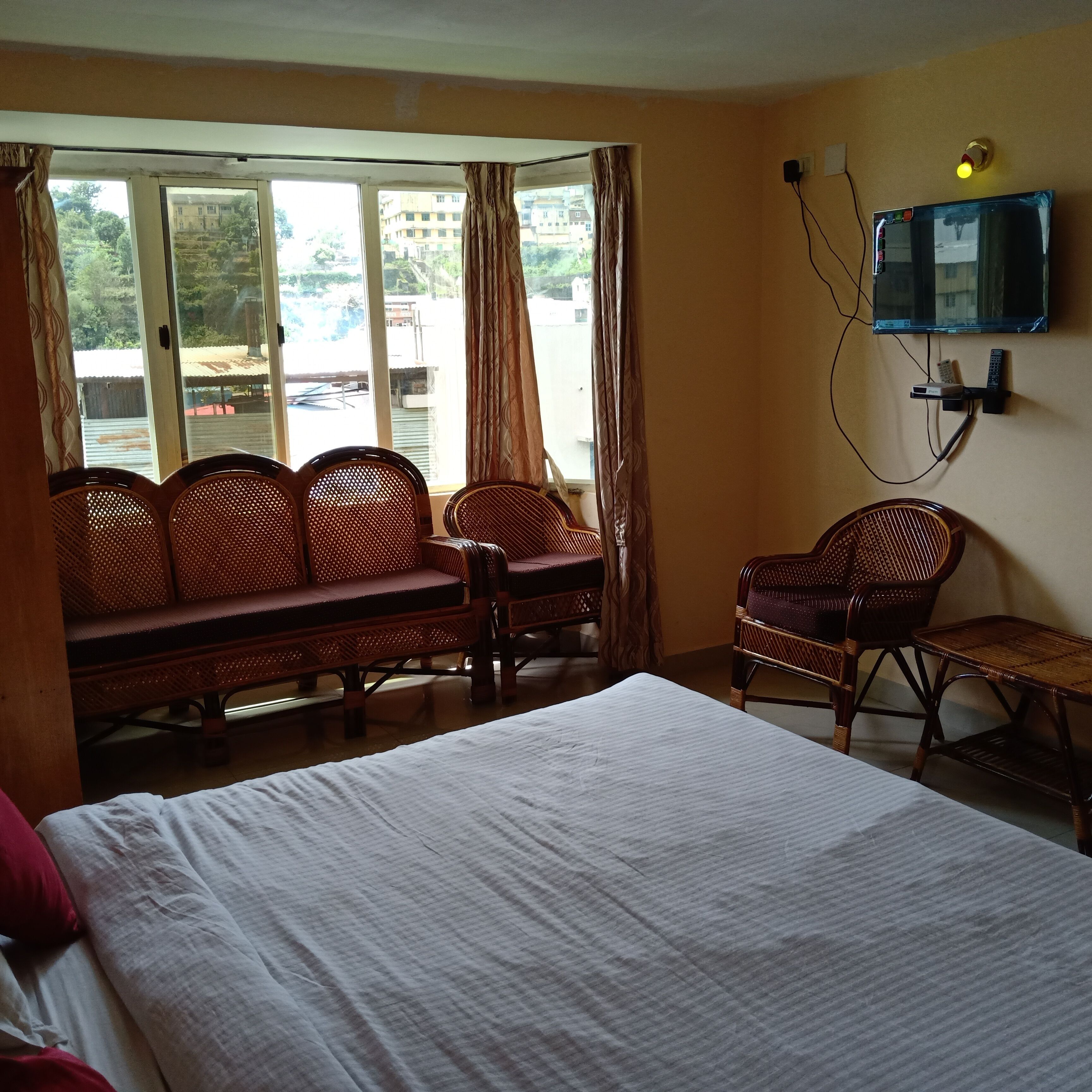 Hotelkamer met een bed, een slaapbank en een stoel. Er is een raam met gordijnen, een aan de muur bevestigde televisie en een stopcontact.