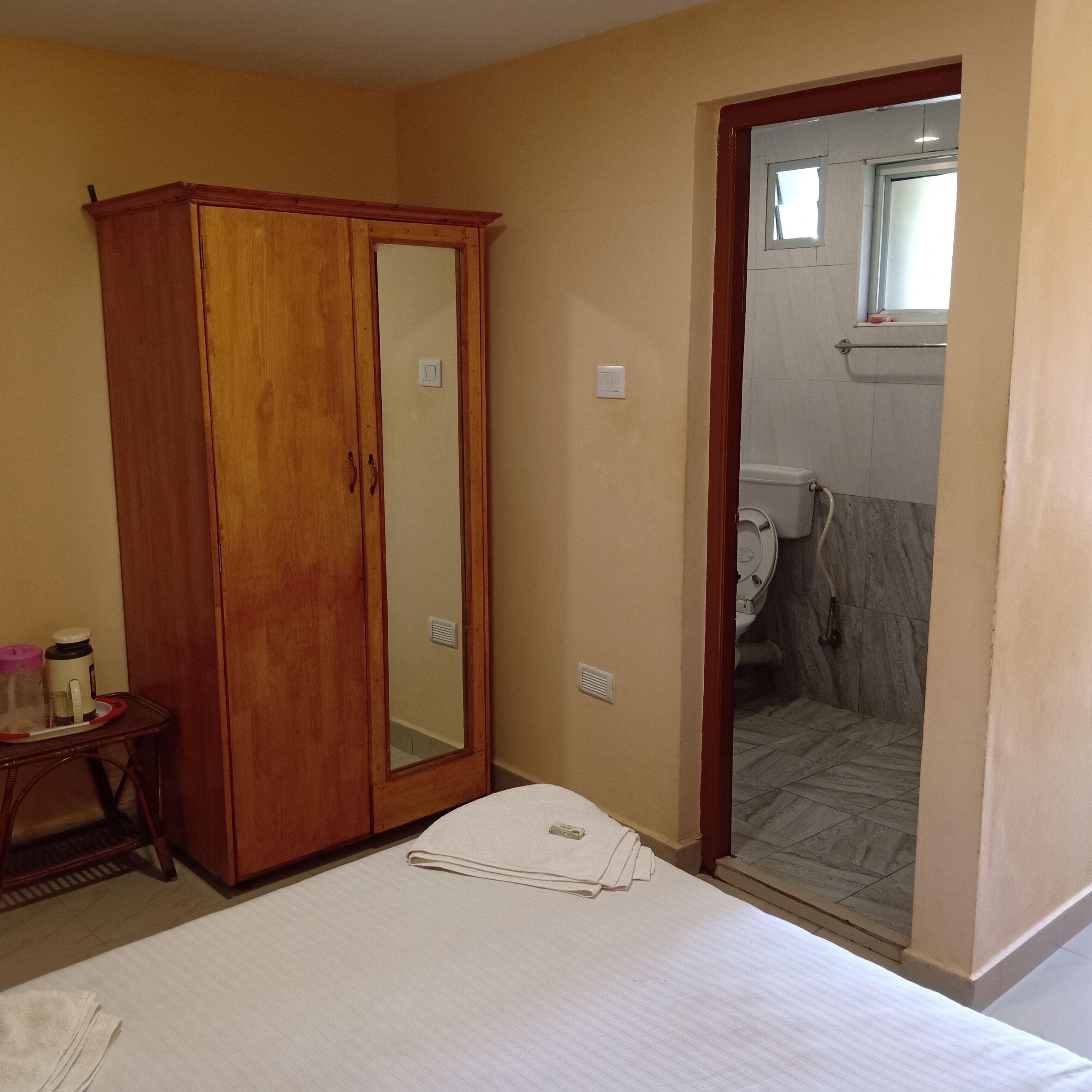 A bedroom with a wooden wardrobe, a small table with a thermos, and a neatly made bed.