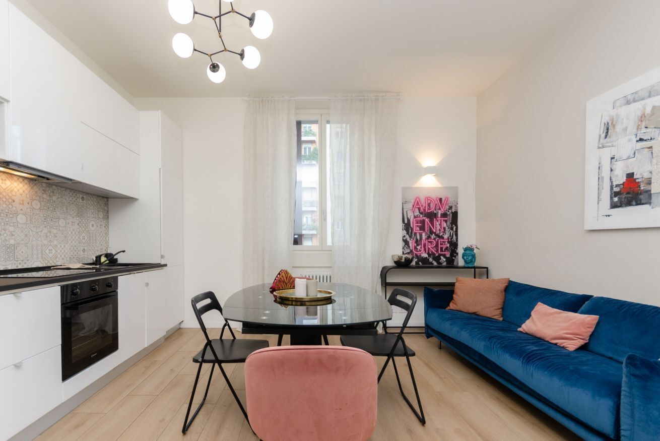Una cocina moderna con armarios blancos, encimera negra y una mesa redonda con sillas. Hay un sofá azul con cojines rosados y un cuadro en la pared.
