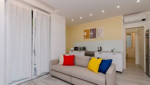 Un salon moderne avec un canapé beige, une porte-fenêtre coulissante, un coin cuisine doté de meubles blancs et un mur orné de trois tableaux encadrés.