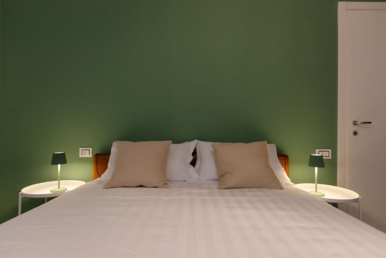 Ein ordentlich bezogenes Bett mit zwei Kissen, ein Nachttisch mit Lampe und eine grüne Wand.