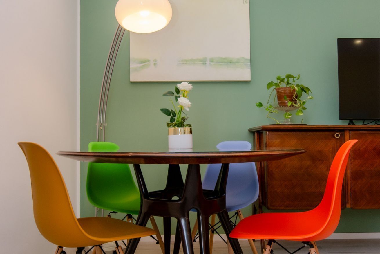 Een eetruimte met een ronde tafel, kleurrijke stoelen, een vloerlamp, een schilderij en een televisie op een kast.