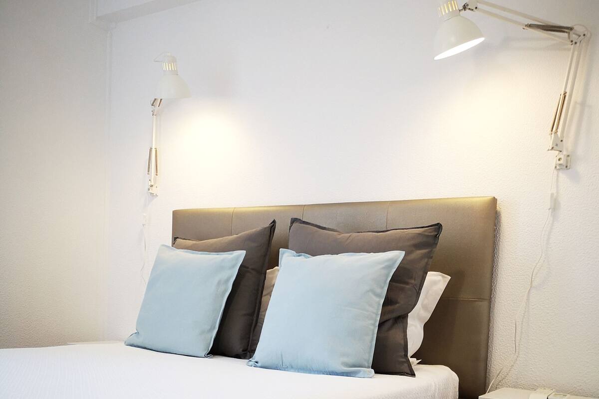 Una cama bien hecha con una colcha blanca y dos almohadas, una mesita de noche con una lámpara y piso de madera.