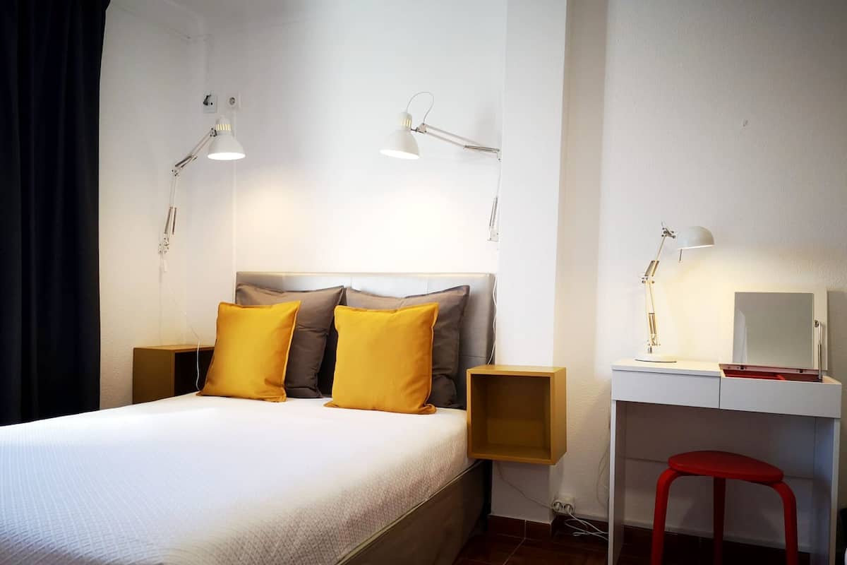 Un dormitorio ordenado con cama, mesitas de noche, un escritorio con silla y un espejo.