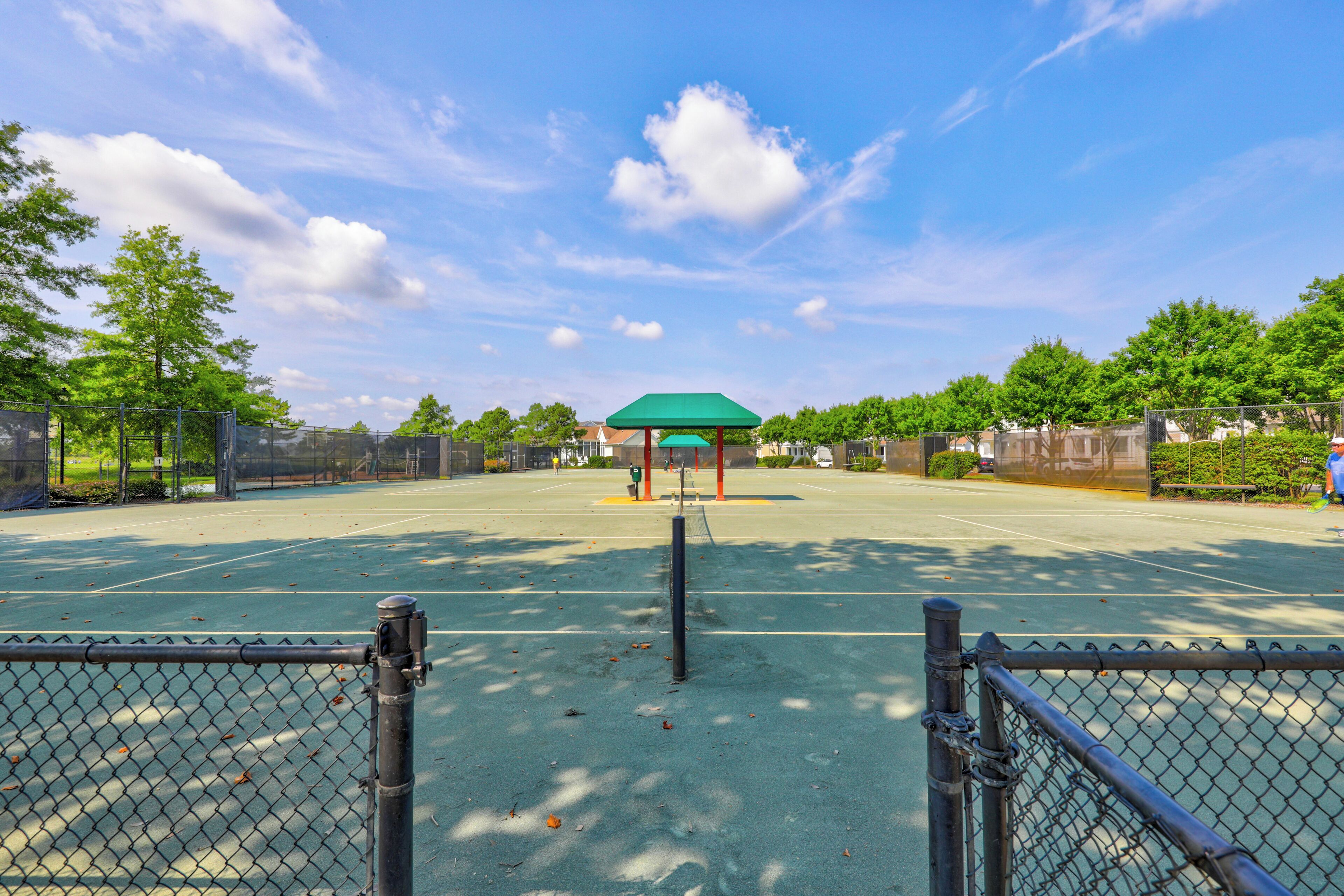An outdoor tennis court with a green-roofed pavilion, surrounded by trees and a clear sky.