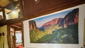 A room with a large landscape painting on the wall, a brick wall, and a wooden ceiling. - A rustic and charming chalet nestled among centuries-old Jatobá trees! (Piraputanga)