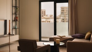 A modern living room with a sofa, armchair, and a small coffee table. A bookshelf with decorative items is against the wall, and a large window offers a view of buildings outside. - Brassbell Zamalek Diplomatic Zone Near Marriott (Cairo)