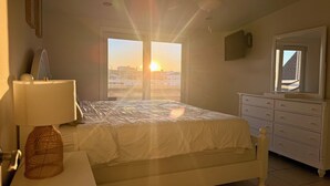 Ein Schlafzimmer mit Bett, Nachttisch mit Lampe, Kommode und Fenster mit Blick auf Gebäude und die Sonne.