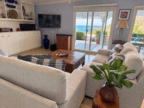 A living room with a sofa, coffee table, and a TV. There is a view of the ocean through large windows. - Seachange--views, pool, AC and generator (Christiansted)