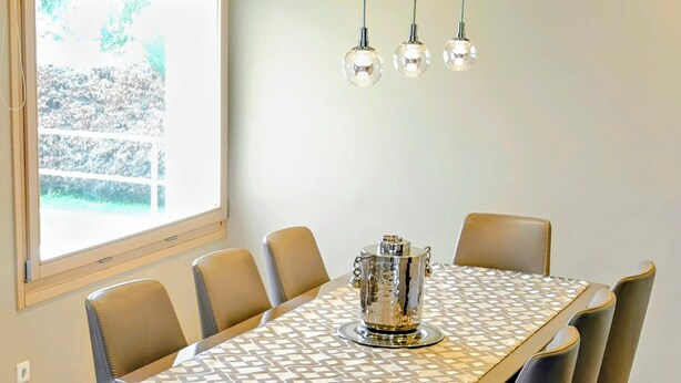 A dining area with a rectangular table, six chairs, a decorative centerpiece, pendant lights, and a window with a view of greenery. - Serene,  spacious 4-bedroom villa in beautiful Pelion, located on the west side (Artemis)