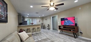 A living room with a sofa, a TV on a wooden stand, a ceiling fan, and a kitchen area in the background.