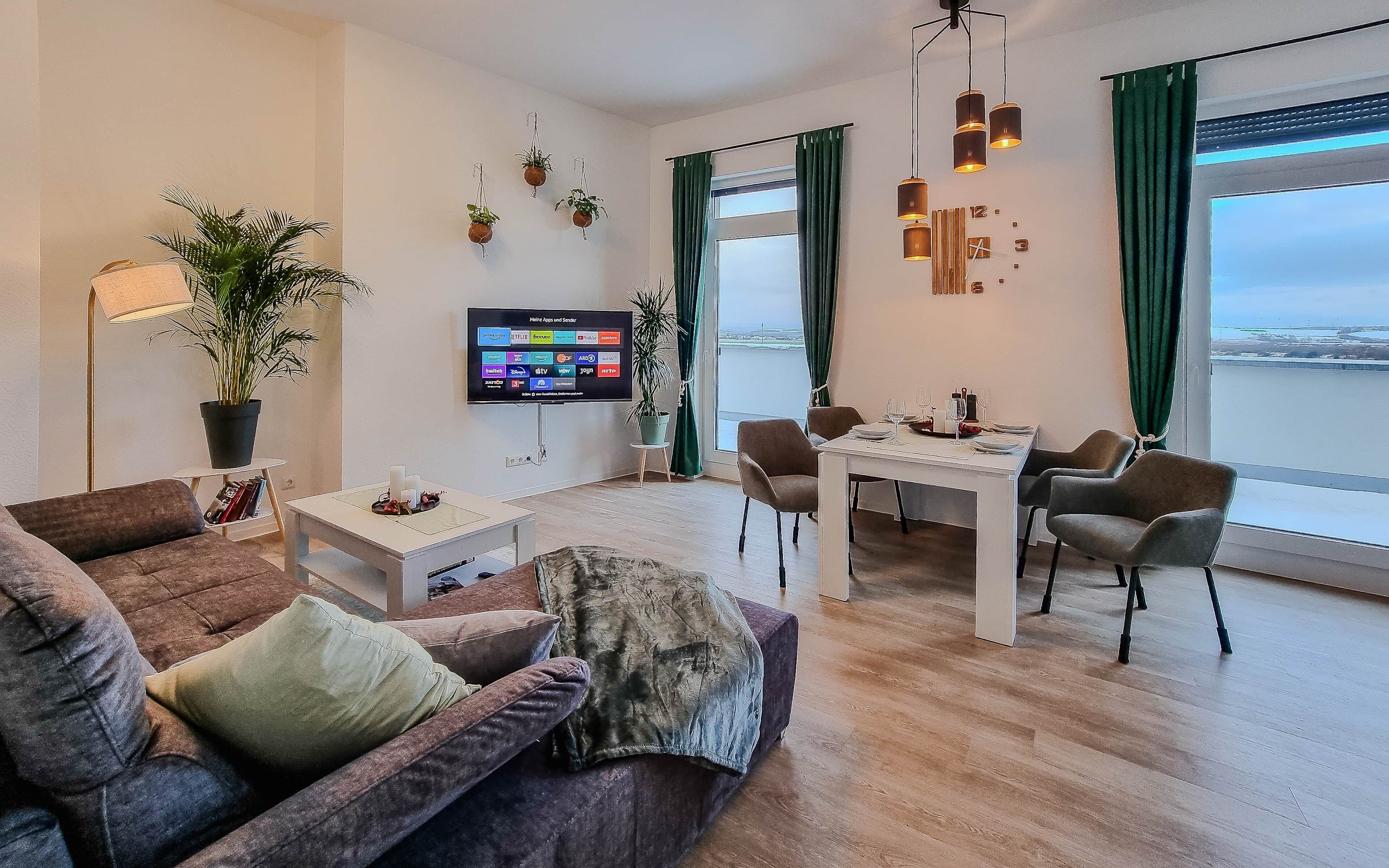 Ein modernes Wohnzimmer mit Sofa, Esstisch für zwei Personen, Wand-Fernseher und großem Fenster mit Vorhängen.