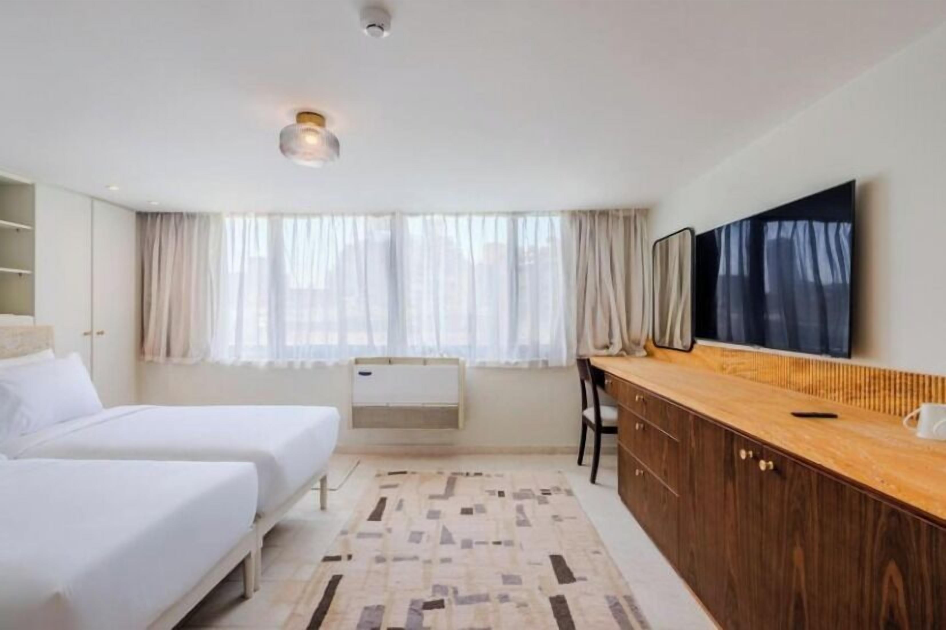 Een hotelkamer met twee bedden, een houten bureau, een flatscreen televisie en een groot raam met dunne gordijnen.