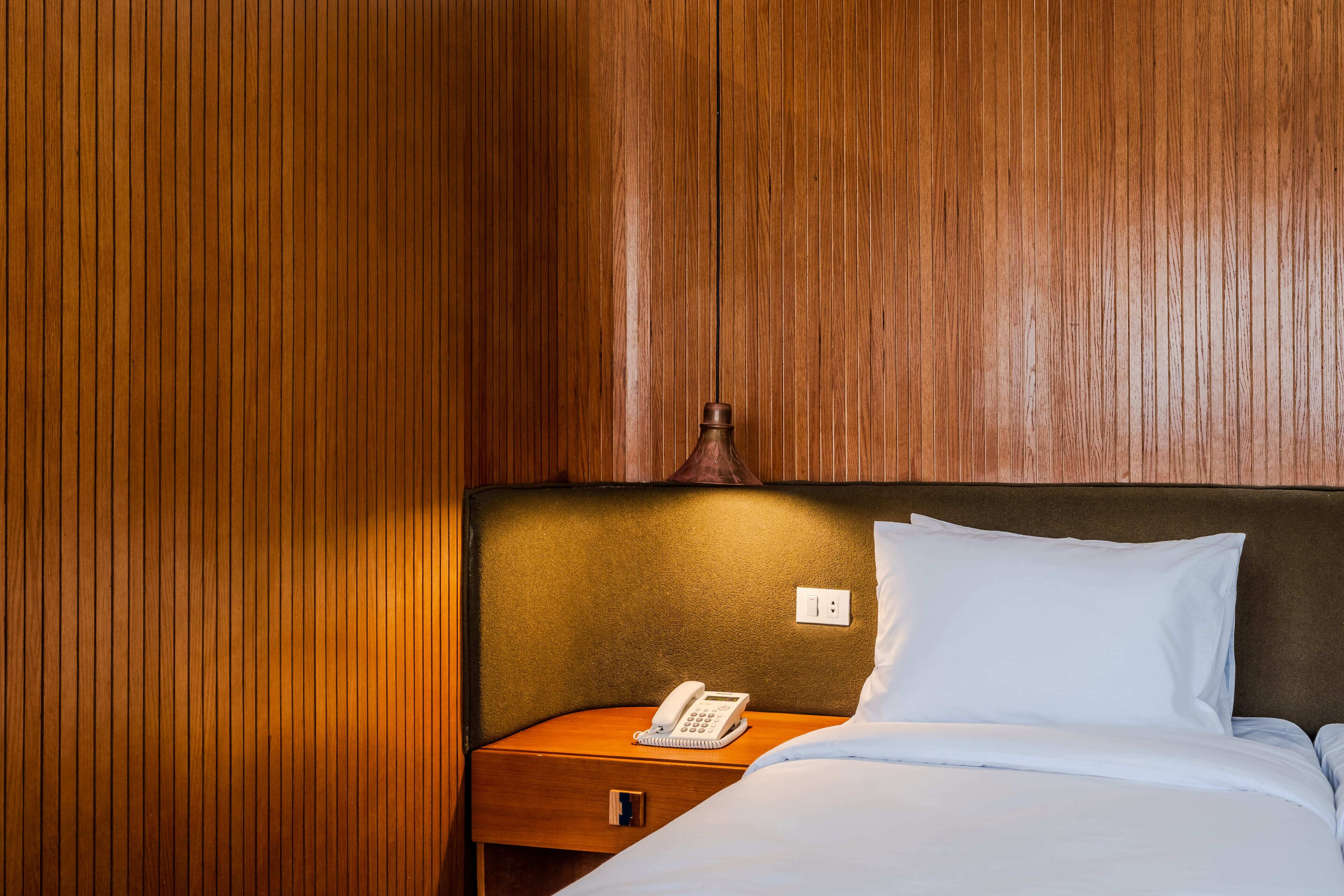 Habitación de hotel con pared de paneles de madera, mesita de noche con teléfono, almohada y cama con ropa blanca.