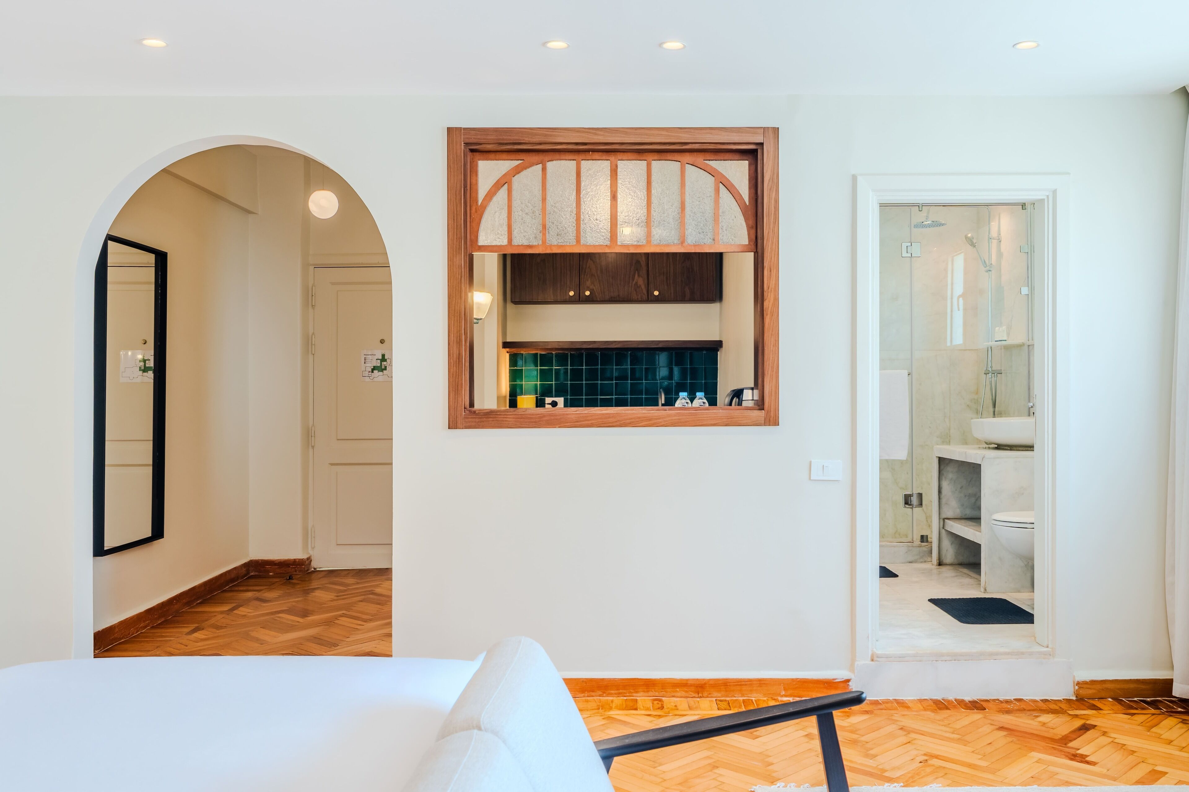 A modern hotel room with a wooden floor, a large bed, a built-in window with a wooden frame, and a separate bathroom with a glass shower enclosure.